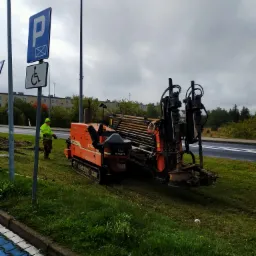 Pomarańczowa maszyna do przewiertów sterowanych Ditch Witch JT2720 Mach 1 na trawniku obok drogi, operator w żółtej kamizelce stoi obok, w tle znak parkingu dla niepełnosprawnych i budynki.