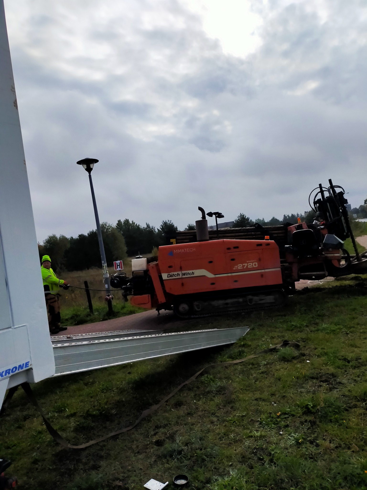 Pomarańczowa maszyna Ditch Witch JT2720 do wierceń horyzontalnych, pracownik w odblaskowej kurtce obsługujący urządzenie na tle pochmurnego nieba i drzew.