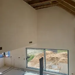 Wnętrze w stanie surowym po nałożeniu tynków na ściany poddasza, widoczne drewniane krokwie dachu, okno z widokiem na hałdę piachu, instalacja elektryczna na podłodze i prowizoryczny stolik...