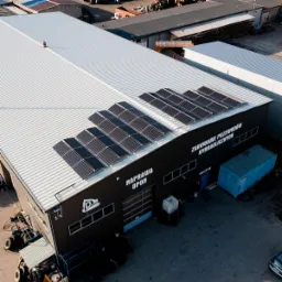 Widok z góry na budynek warsztatu z panelami słonecznymi zamontowanymi na dachu, z napisem 'Naprawa Opon' i 'Zakłuwanie Przewodów Hydraulicznych' na ścianie. Obok budynku kontener i sprzęt...