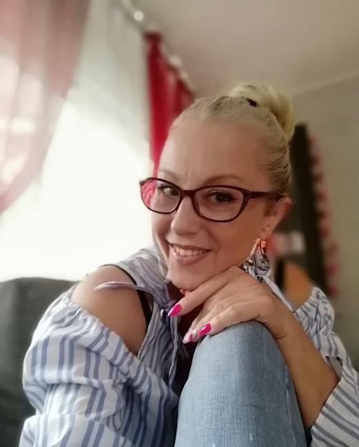 Portret uśmiechniętej kobiety w okularach, z upiętymi włosami, w bluzce w paski, siedzącej na kanapie z kolanami pod brodą, z widocznym manicure w kolorze fuksji.