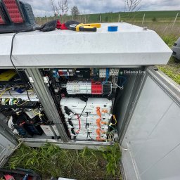Zielona Era - Otwarta szafa sterownicza z panelami i okablowaniem, narzędzia na zewnątrz, trawa wokół, widok na zielone pole w tle, pochmurne niebo.