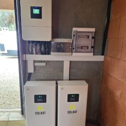 Zielona Era - Dwa białe magazyny energii SOLBAT na kółkach, obok ściany z cegły, z podłączonym inwerterem i rozdzielnicą. Widoczne okablowanie i elementy instalacji.