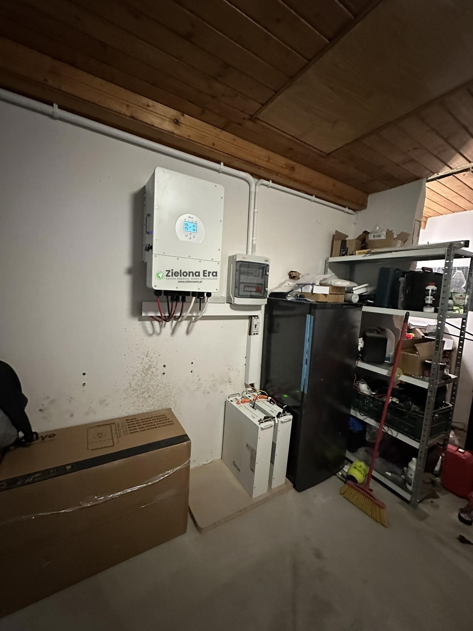 Wnętrze pomieszczenia z zamontowanym inwerterem firmy Zielona Era, licznikiem energii elektrycznej, magazynem energii oraz regałem z różnymi przedmiotami.