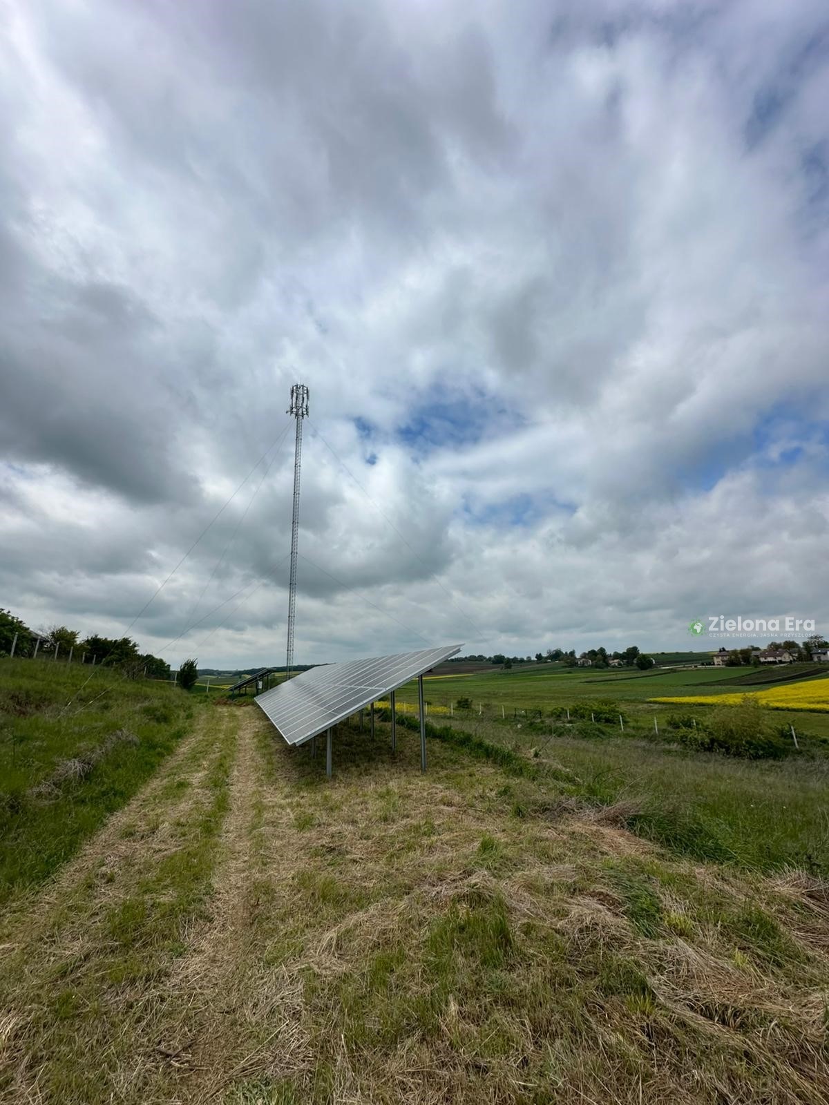 Panel słoneczny na łące z wieżą telekomunikacyjną w tle, pochmurne niebo. Instalacja na nierównym terenie, widok z dołu, podkreślający perspektywę.