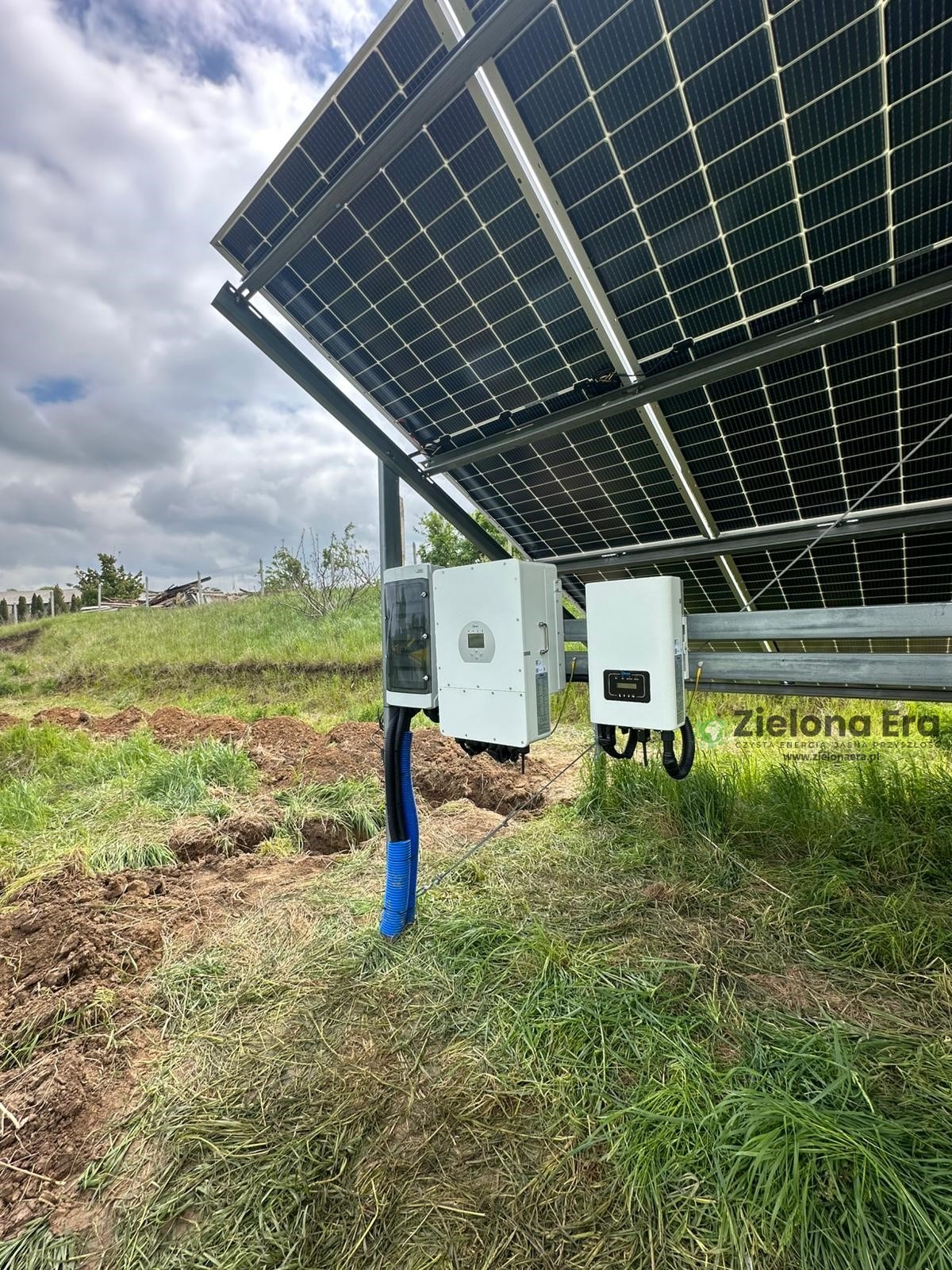 Instalacja paneli słonecznych z inwerterami na trawiastym terenie, widoczne kable w niebieskiej osłonie i fragment krajobrazu w tle.