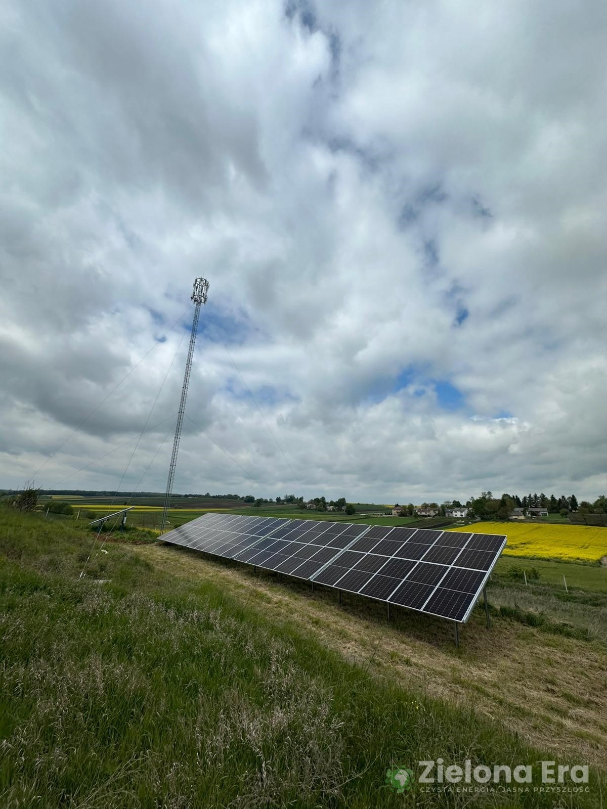 Panel słoneczny na wzgórzu z wieżą telekomunikacyjną w tle, pola rzepaku i zachmurzone niebo. Widoczny logotyp 'Zielona Era'.
