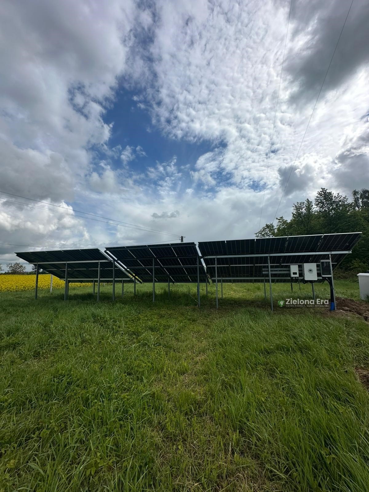 Instalacja paneli na gruncie, widok z dołu na konstrukcję i falowniki, w tle pole rzepaku i pochmurne niebo. Znak wodny w prawym dolnym rogu.