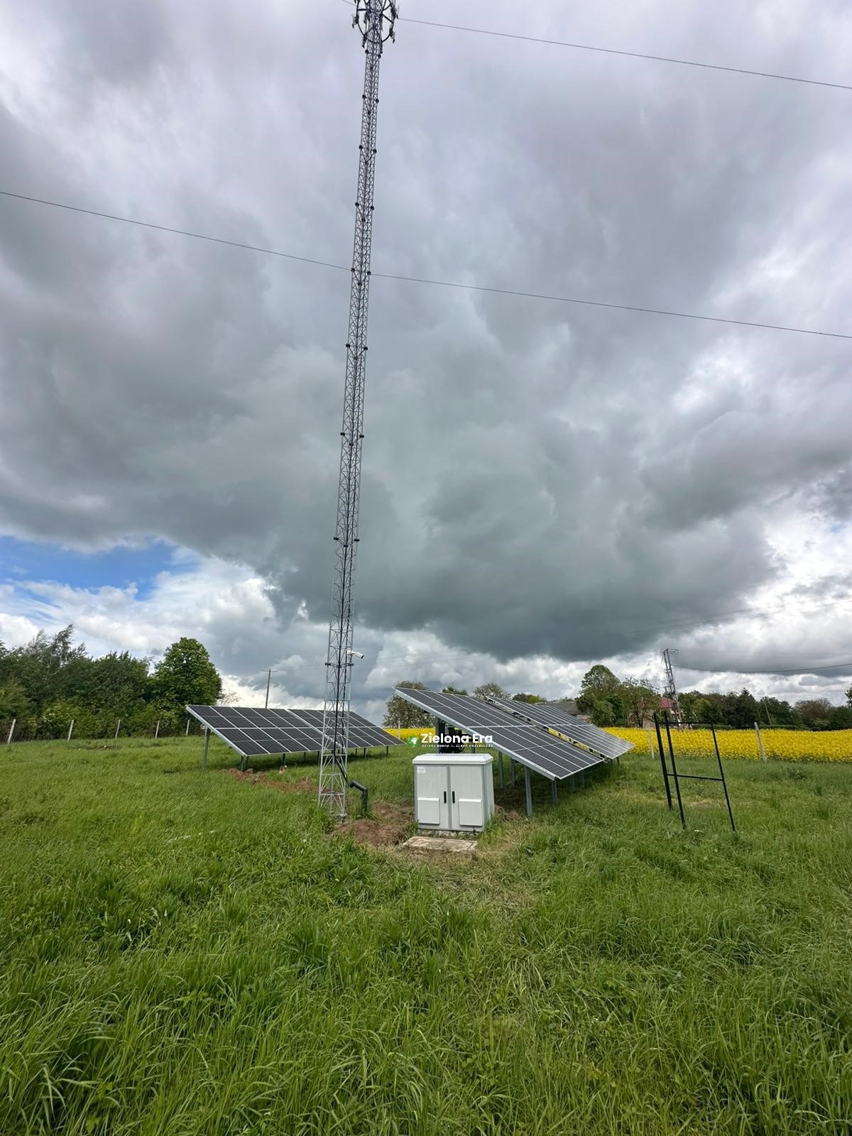 Instalacja paneli słonecznych na łące obok wieży telekomunikacyjnej, pod pochmurnym niebem. Widoczne żółte pole w tle i szafka sterownicza.