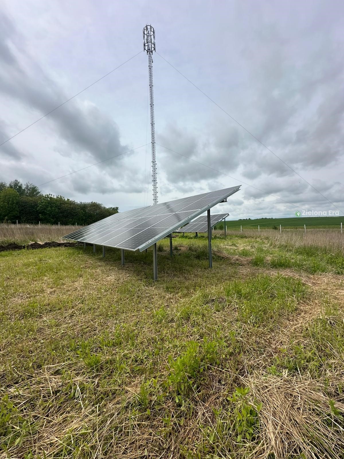 Instalacja paneli słonecznych na łące, z wieżą telekomunikacyjną w tle i pochmurnym niebem. Widok na panele pod kątem.