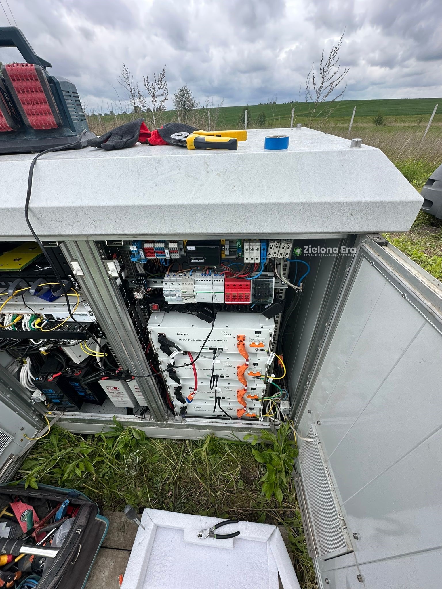 Otwarta szafa sterownicza z panelami i okablowaniem, narzędzia na zewnątrz, trawa wokół, widok na zielone pole w tle, pochmurne niebo.