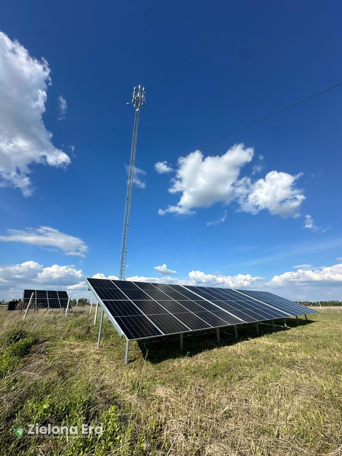 Instalacja paneli słonecznych na łące, w tle wieża telekomunikacyjna. Błękitne niebo z chmurami. Panele ustawione pod kątem, widoczne metalowe konstrukcje.