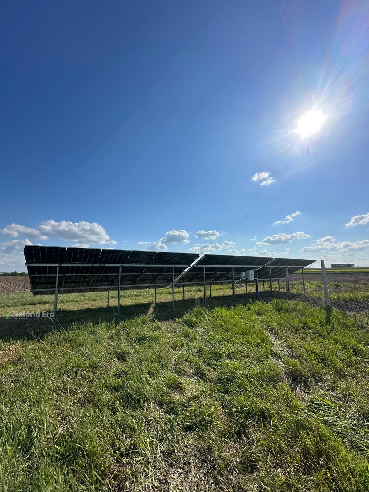 Instalacja paneli na łące, widok z dołu, błękitne niebo z chmurami, słoneczny dzień. Panele zamontowane na metalowej konstrukcji.