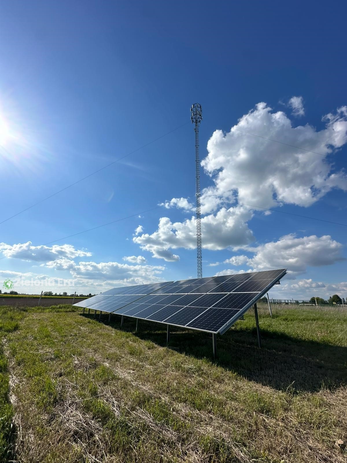 Instalacja paneli słonecznych na łące, w tle wieża telekomunikacyjna i błękitne niebo z chmurami. Widok z dołu, podkreślający przestrzeń i nowoczesność.