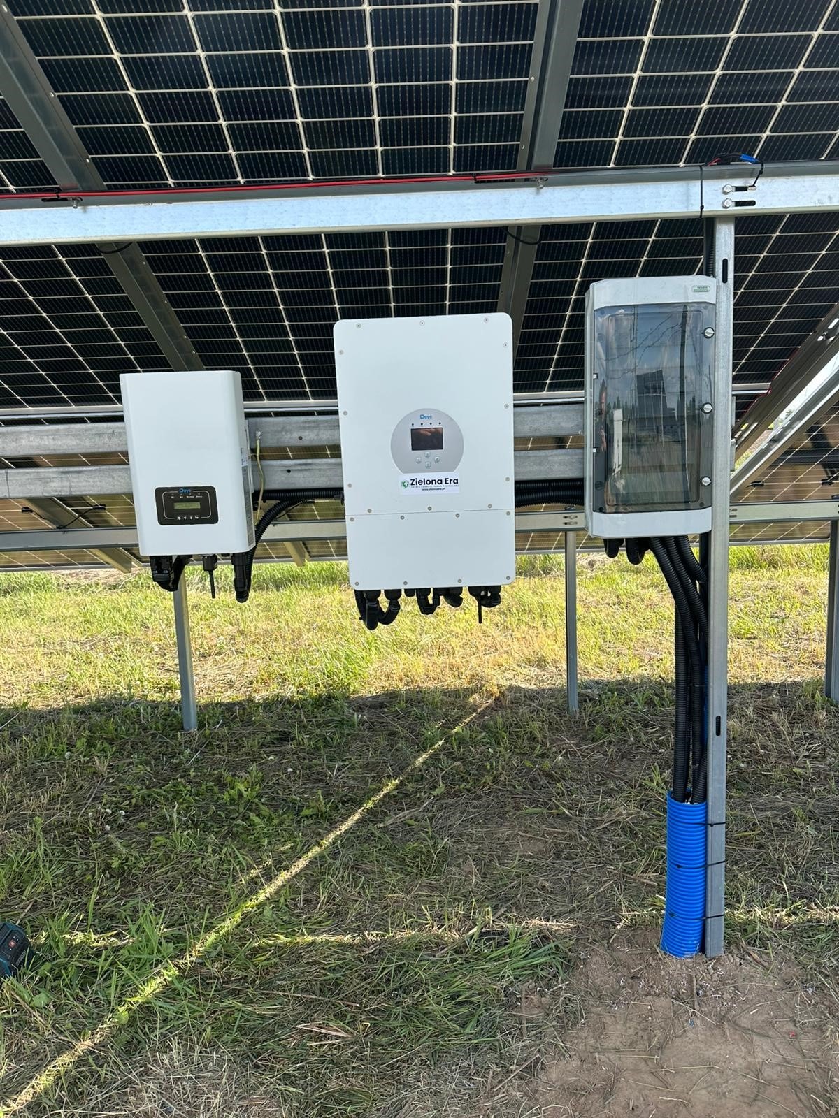 Instalacja z panelami słonecznymi, widoczne inwertery i zabezpieczenia w obudowie. Kable w peszlach. Montaż na trawiastym terenie.