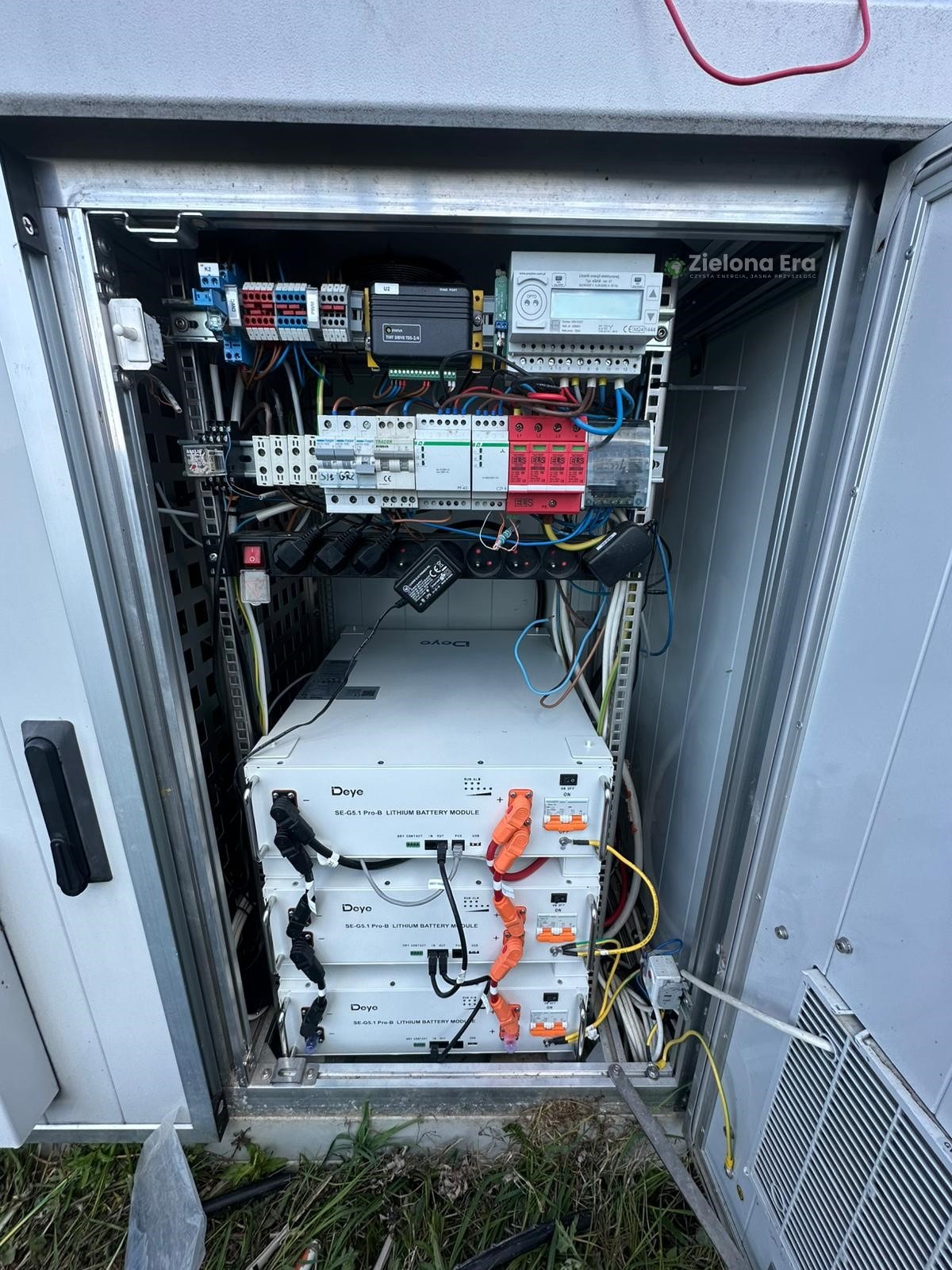Otwarta szafa sterownicza z trzema modułami bateryjnymi Deye SE-G5.1 Pro-B, licznikiem energii i zabezpieczeniami. Widoczne okablowanie i elementy instalacji elektrycznej.