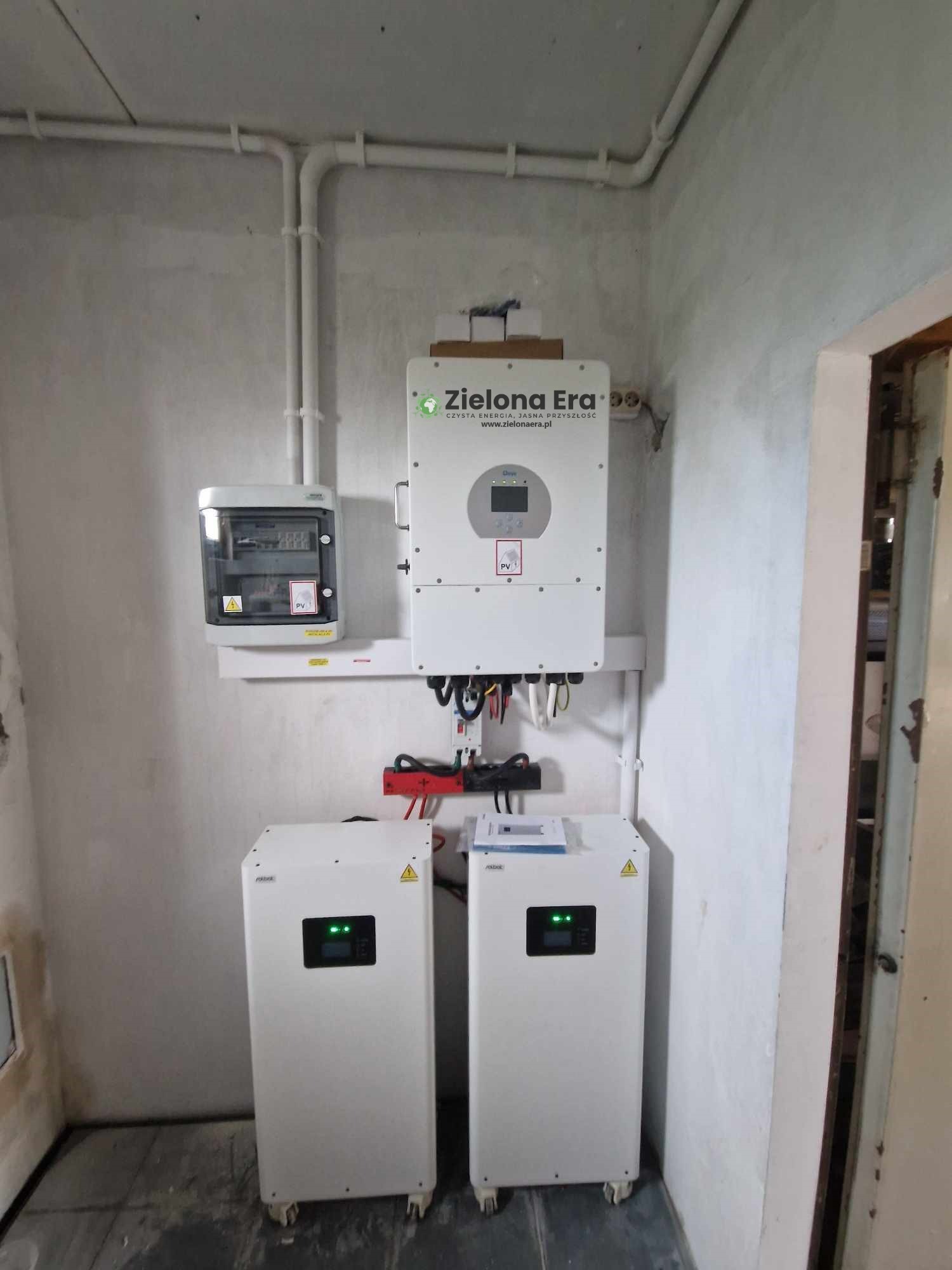 Kompletna instalacja fotowoltaiczna w pomieszczeniu: falownik, rozdzielnica i dwa magazyny energii na kółkach. Widoczne okablowanie i elementy montażowe.