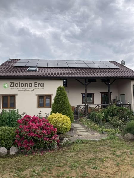 Panele słoneczne na dachu domu z brązową dachówką, widok z ogrodu z kwitnącymi krzewami. Logo firmy na ścianie budynku.