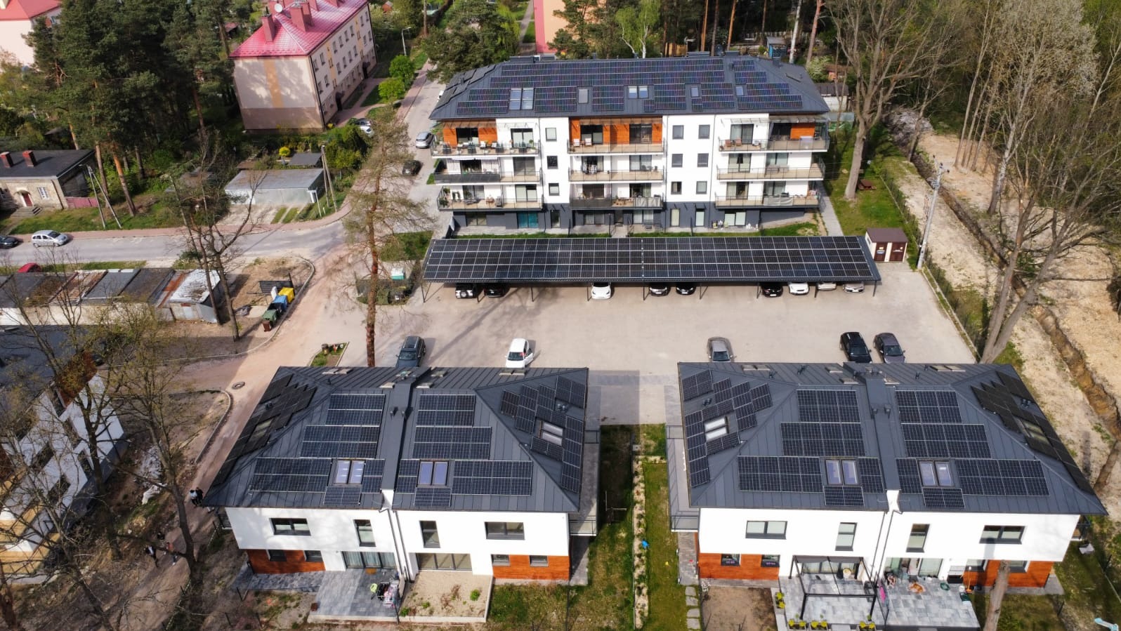 Widok z drona na budynki z panelami słonecznymi na dachach i wiatą parkingową. Kompleks mieszkalny z instalacją OZE w słoneczny dzień.