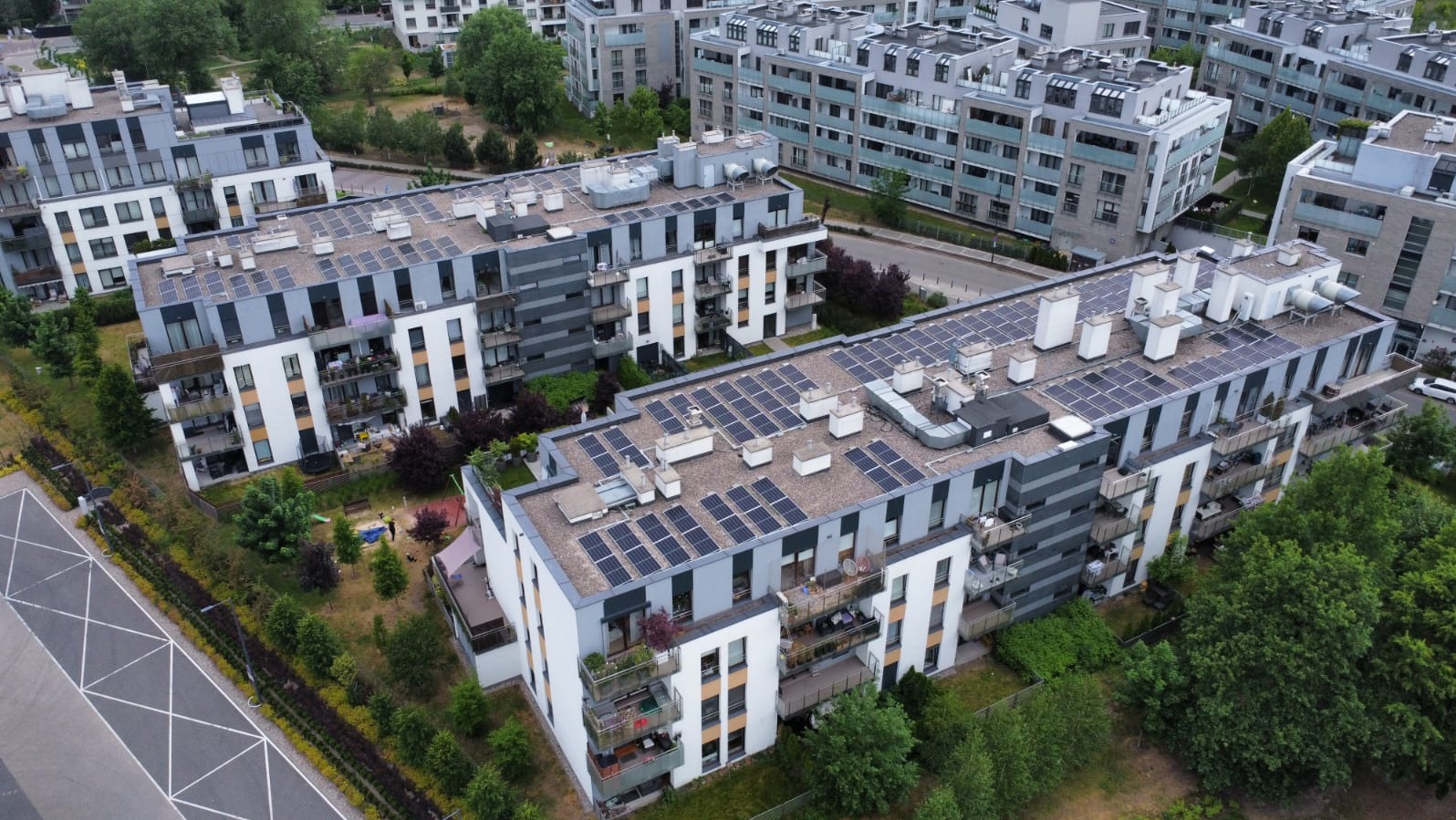 Widok z góry na nowoczesne budynki mieszkalne z panelami słonecznymi na dachach, otoczone zielenią i innymi budynkami.