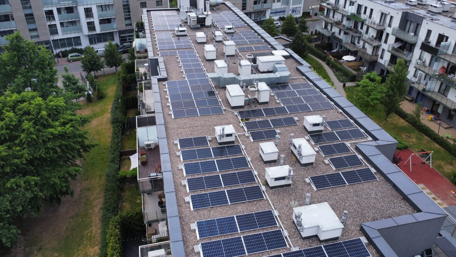 Widok z góry na dach budynku z panelami słonecznymi i klimatyzatorami, otoczony zielenią i budynkami mieszkalnymi. Ujęcie z drona.