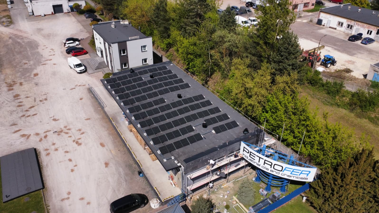 Widok z góry na budynek z panelami słonecznymi na dachu, z logo firmy Petrofer. W tle drzewa i inne budynki, na placu zaparkowane samochody.