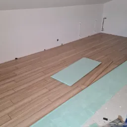 Proces układania paneli podłogowych w pomieszczeniu z białymi ścianami, widoczna zielona podkładka izolacyjna i panele imitujące drewno.