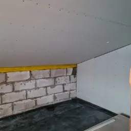 Poddasze w trakcie prac wykończeniowych: ściany z płyt kartonowo-gipsowych, surowy mur z żółtą folią zabezpieczającą, czarna folia na podłodze, widoczne wkręty w suficie.