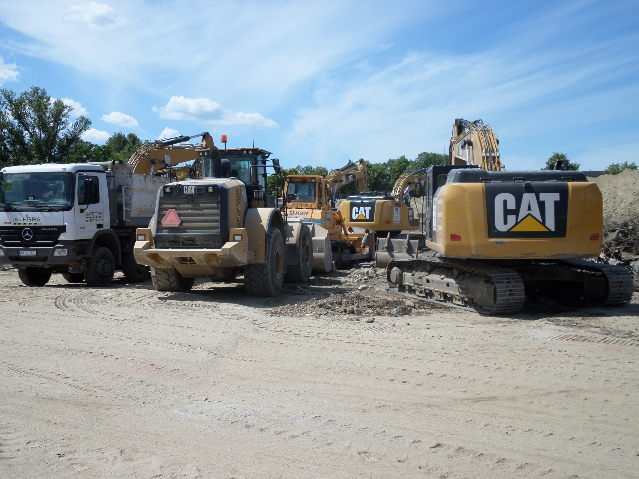 Szeroki kadr przedstawia zaparkowane obok siebie maszyny budowlane: wywrotkę Mercedes Actros, ładowarkę kołową CAT, koparkę HSW oraz dwie koparki gąsienicowe CAT, na piaszczystym terenie placu...