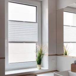 Biała, podwójna plisa okienna w łazience z białymi płytkami, częściowo zasłonięta, z dekoracyjną trawą w złotej doniczce na parapecie.