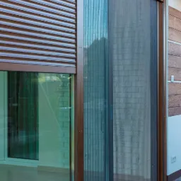 Zewnętrzna moskitiera plisowana w kolorze brązowym, zamontowana na tarasie, widoczna obok opuszczonej rolety zewnętrznej, z elementami muru z cegły w tle.