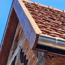 Fragment odnowionego dachu z terakoty, z widoczną drewnianą konstrukcją i nową metalową rynną, na tle błękitnego nieba.