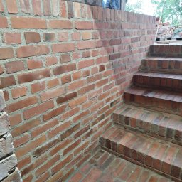 Revita GmbH - Murowana ściana i schody z czerwonej cegły, widoczna konstrukcja drewniana i fragment osoby w tle, prawdopodobnie w trakcie budowy lub remontu.