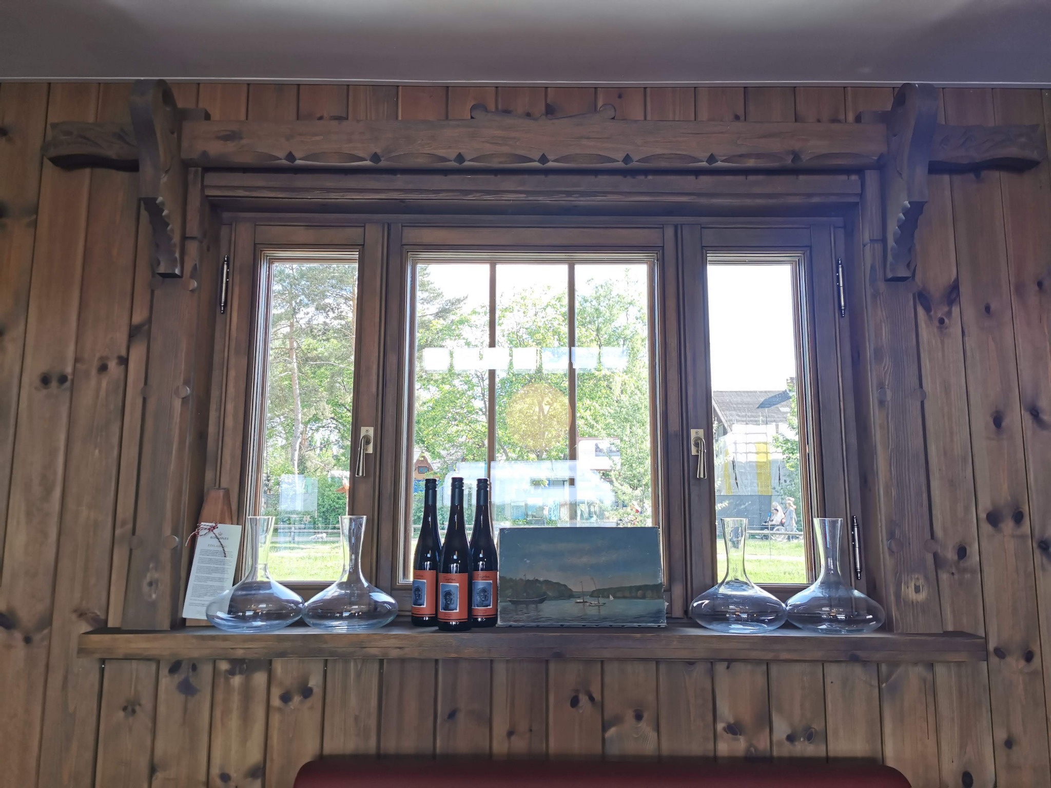 Drewniane okno z dekoracyjnymi zdobieniami, na parapecie karafki, butelki wina i obraz z motywem morskim, ściany wykończone drewnianą boazerią.