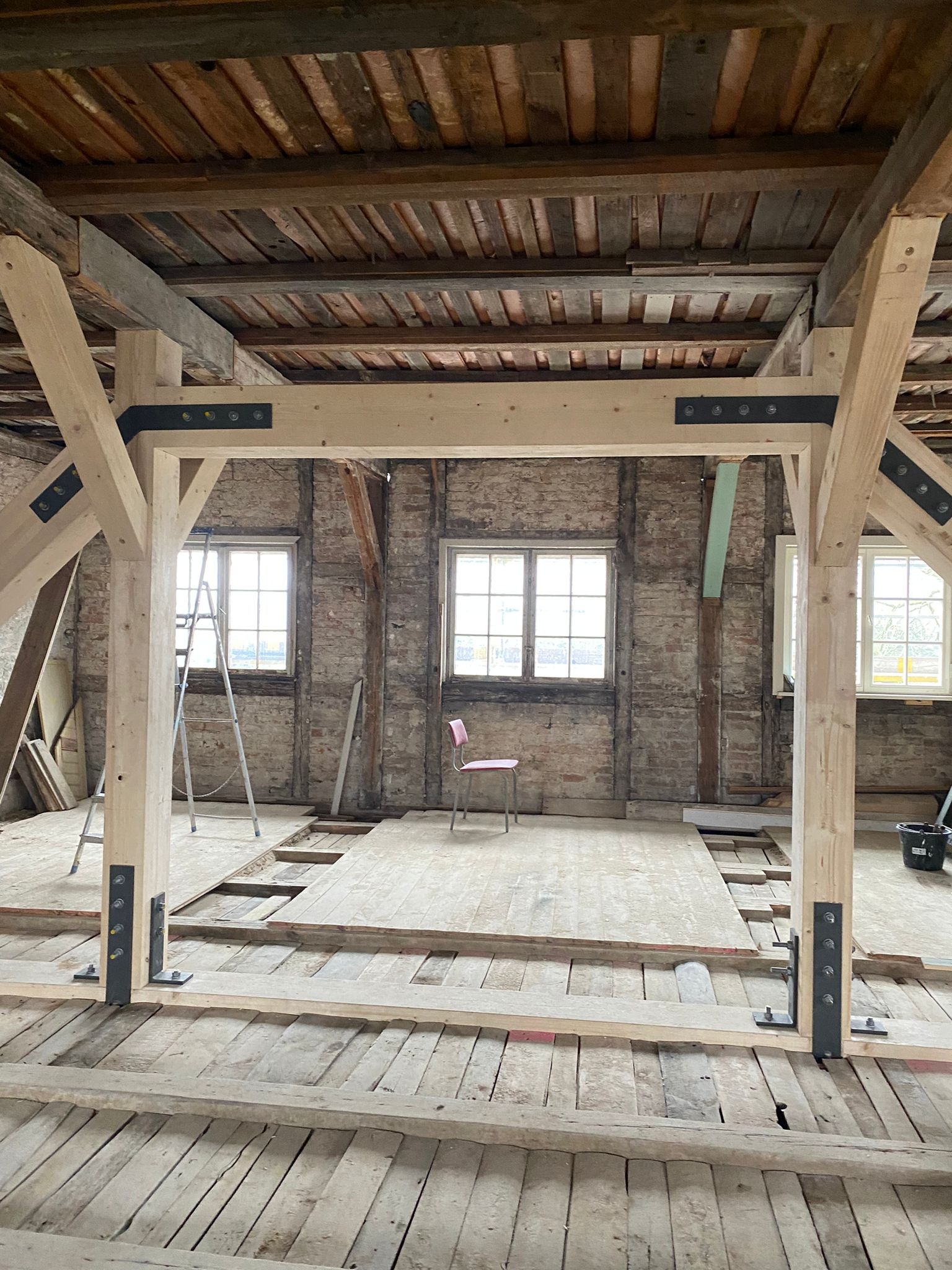 Wnętrze poddasza w trakcie remontu z odsłoniętą cegłą, drewnianą konstrukcją nośną wzmocnioną metalowymi elementami, częściowo ułożoną podłogą i różowym krzesłem w tle.