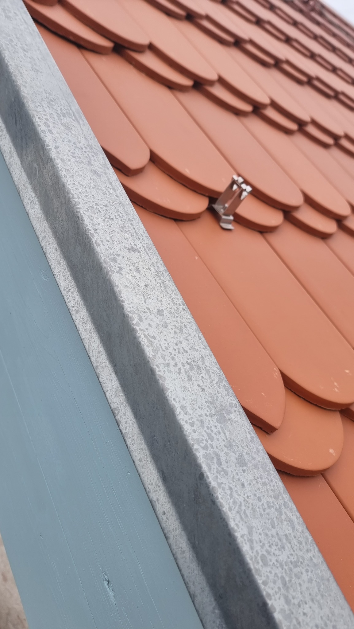 Fragment dachu z ceramiczną dachówką karpiówką w kolorze ceglastym, z widocznym metalowym zabezpieczeniem przeciwśniegowym i szarą obróbką blacharską oraz fragmentem pomalowanej na niebiesko deski...