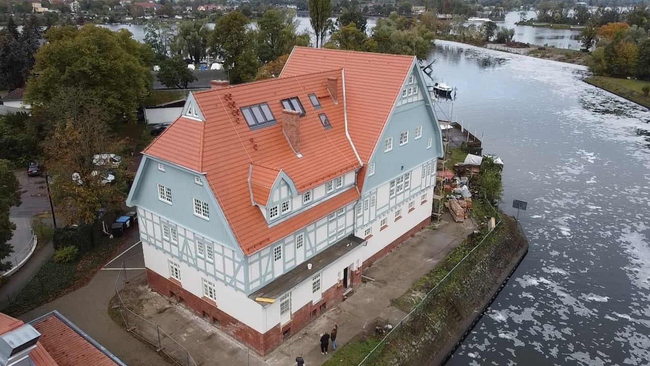Dwupiętrowy budynek z muru pruskiego w trakcie renowacji, z pomarańczową dachówką i oknami dachowymi, położony nad brzegiem rzeki.