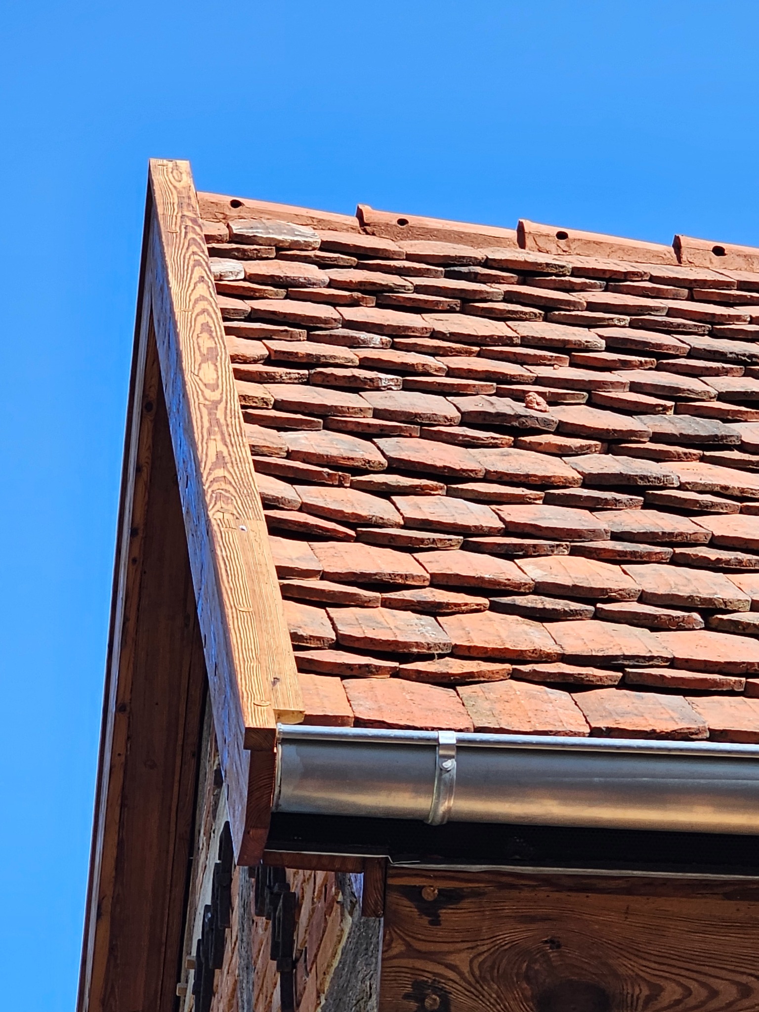 Ceglana dachówka na dachu budynku z drewnianą obróbką krawędzi i metalową rynną, widok z dołu na tle błękitnego nieba.