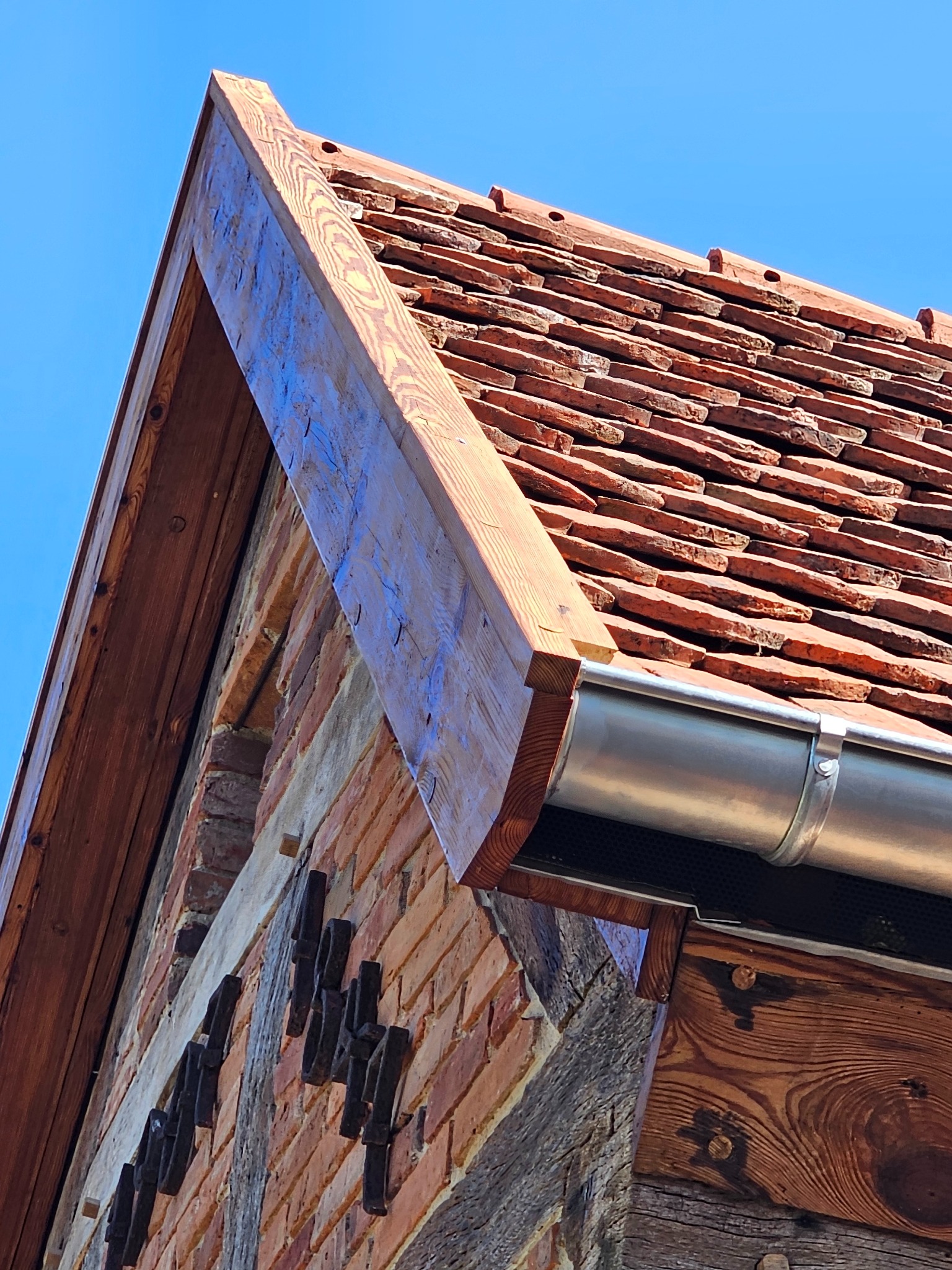 Fragment odnowionego dachu z terakoty, z widoczną drewnianą konstrukcją i nową metalową rynną, na tle błękitnego nieba.