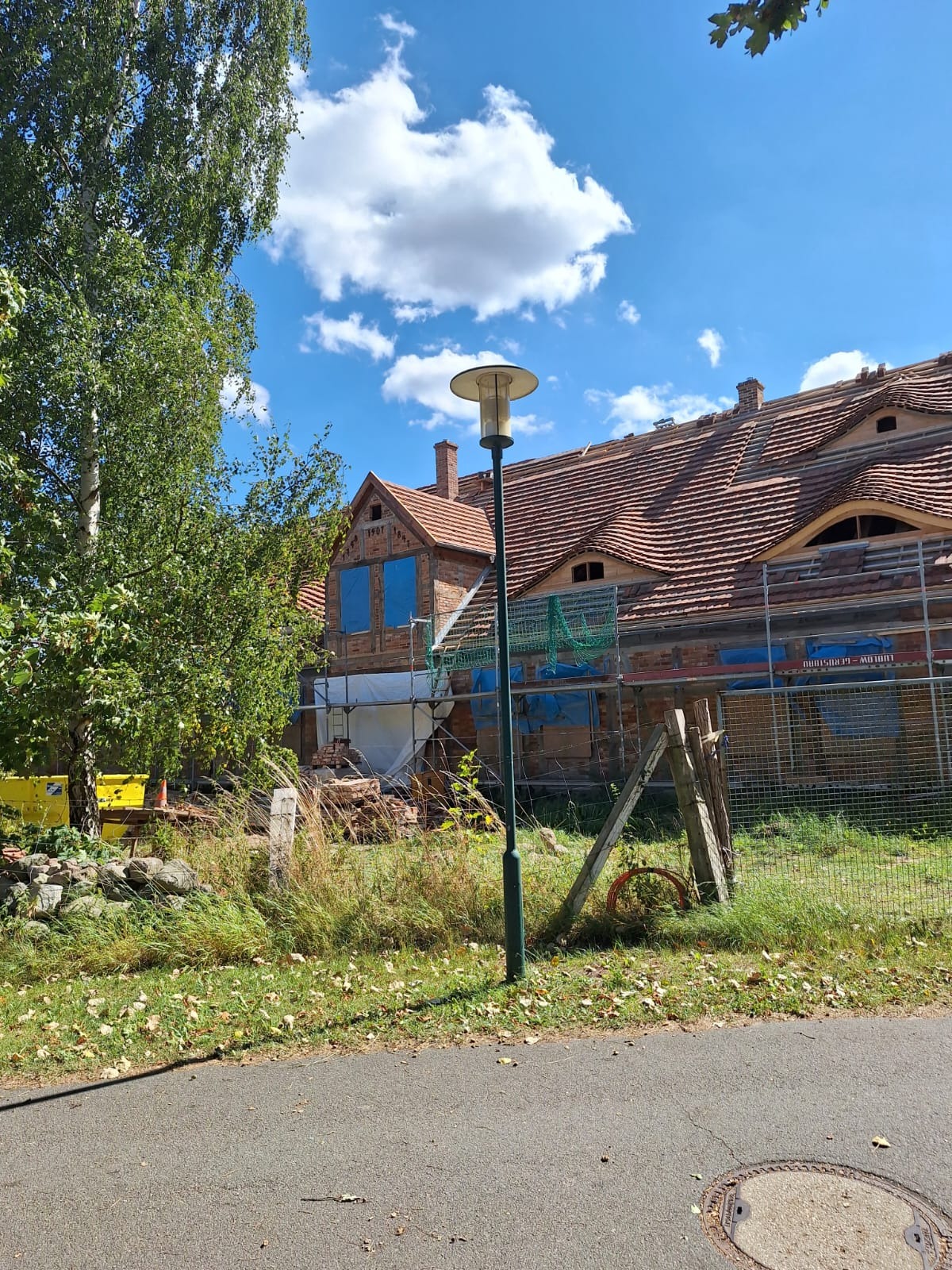 Renowacja ceglanego domu z czerwonym dachem, widoczne rusztowania, zabezpieczone okna, lampa uliczna i fragment chodnika.