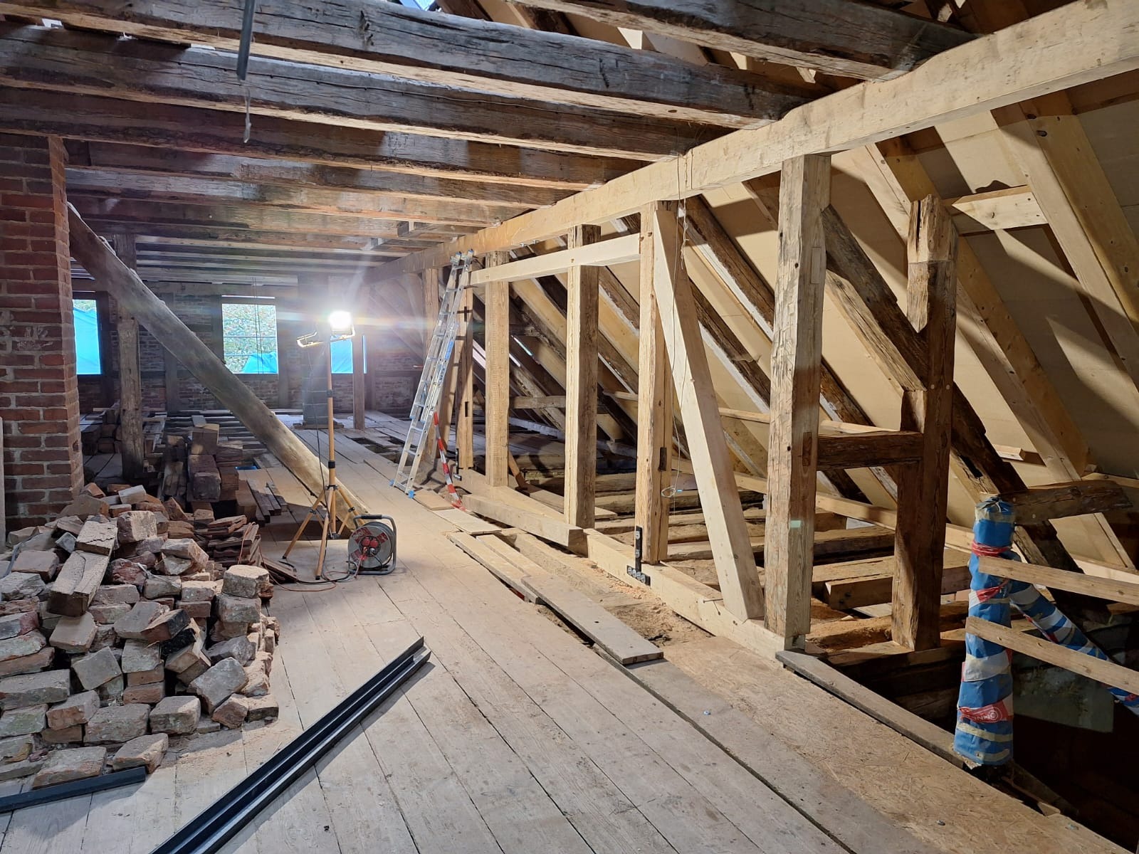 Poddasze w trakcie przebudowy z widoczną konstrukcją drewnianą, stertą cegieł, oknami zabezpieczonymi folią i oświetleniem budowlanym.