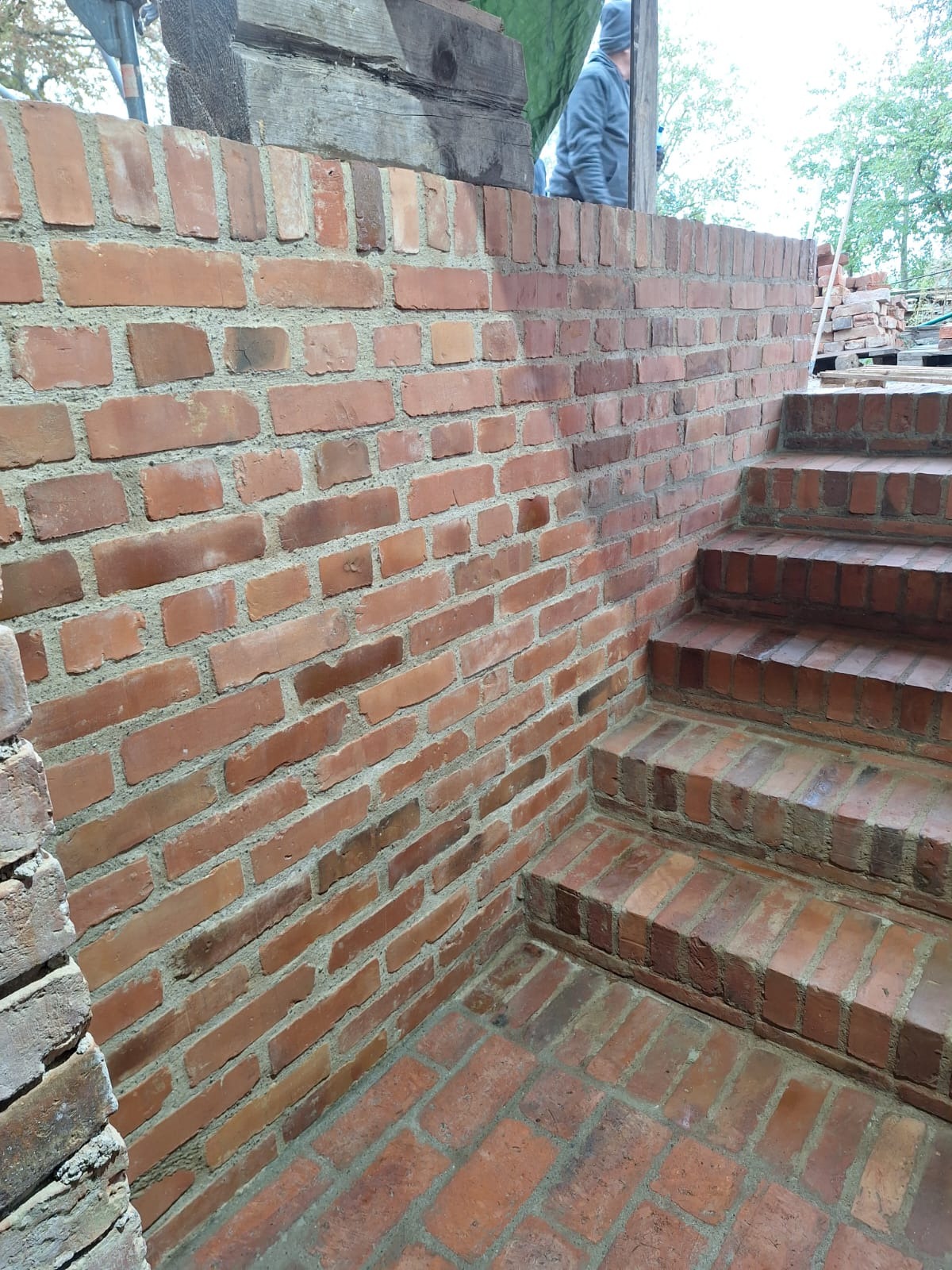 Murowana ściana i schody z czerwonej cegły, widoczna konstrukcja drewniana i fragment osoby w tle, prawdopodobnie w trakcie budowy lub remontu.
