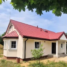 Parterowy dom z keramzytu z czerwonym dachem i białymi oknami, widok z zewnątrz w słoneczny dzień, zarośnięta trawa przed domem.