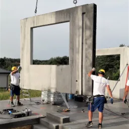 Montaż prefabrykowanej ściany z oknem na placu budowy, robotnicy w kaskach, widoczne łańcuchy podtrzymujące element, na podłożu narzędzia i materiały budowlane.