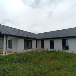 Eurodomy.pl sp. z o.o. - Parterowy dom z keramzytu z ciemnym dachem na tle pochmurnego nieba. Widoczne czarne okna i fragment zadaszonego tarasu. Działka zarośnięta trawą.