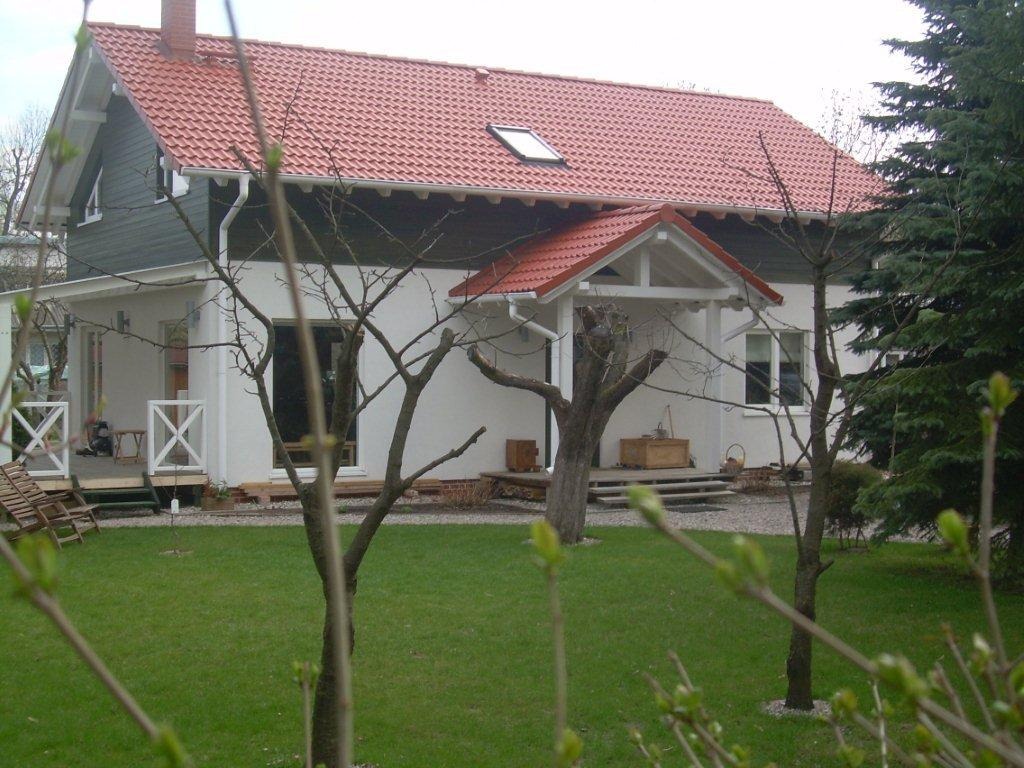 Parterowy dom z czerwonym dachem z dachówki, widok z ogrodu, z drzewami na pierwszym planie i białymi ścianami z szarymi elementami wykończeniowymi.