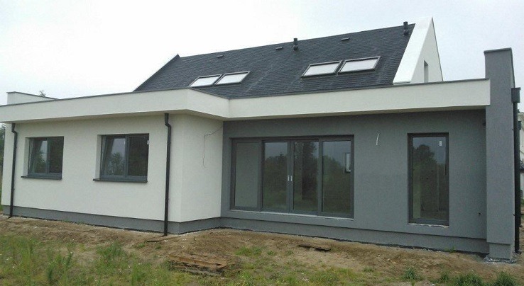 Niski, parterowy dom z płaskim dachem i oknami dachowymi, część elewacji w kolorze białym, część w odcieniach szarości, widoczne okna i przesuwne drzwi tarasowe.