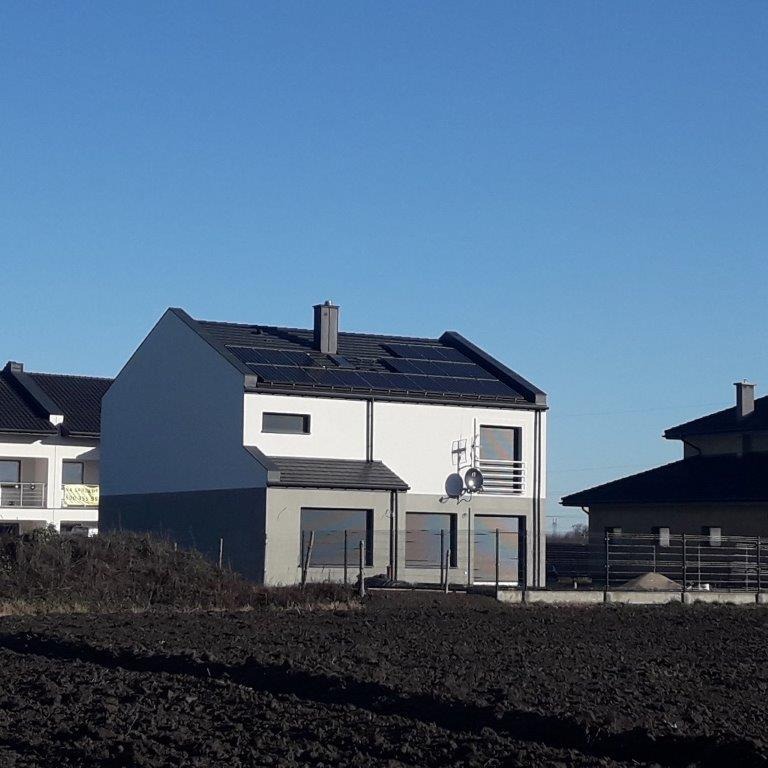 Dwukondygnacyjny dom z ciemną dachówką i panelami słonecznymi, szary cokół, antena satelitarna na elewacji, zaorane pole na pierwszym planie.