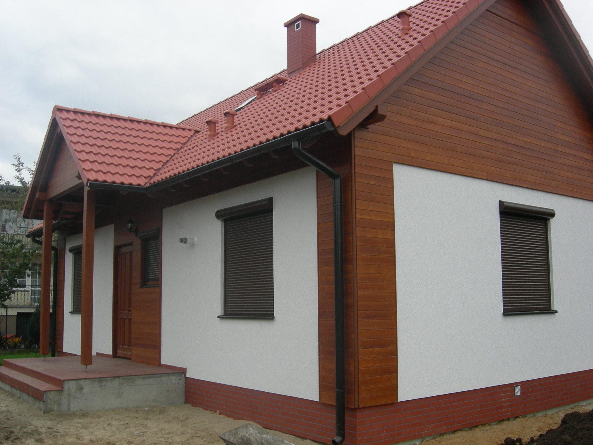 Gotowy dom jednorodzinny z czerwoną dachówką, drewnianą elewacją i roletami zewnętrznymi, widok z boku na tle pochmurnego nieba.