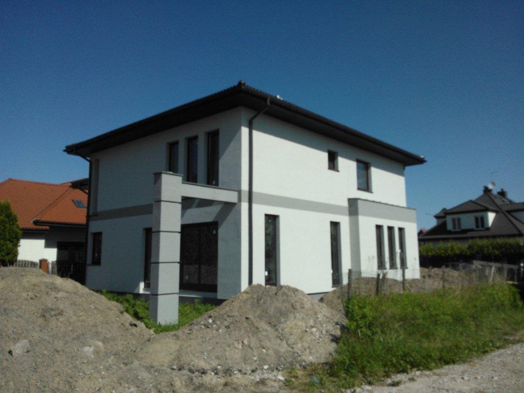 Nowoczesny, dwukondygnacyjny dom w trakcie budowy, z widocznymi stertami ziemi i świeżą zielenią w otoczeniu, na tle błękitnego nieba.